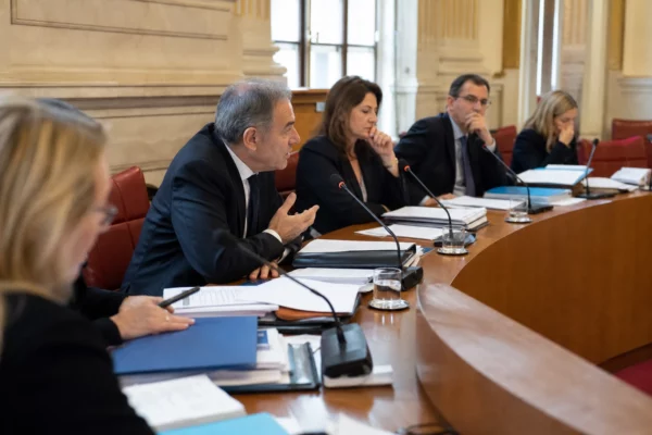 Réunion de commission au Sénat avec micros, dossiers ouverts et silhouettes anonymes en salle parlementaire.