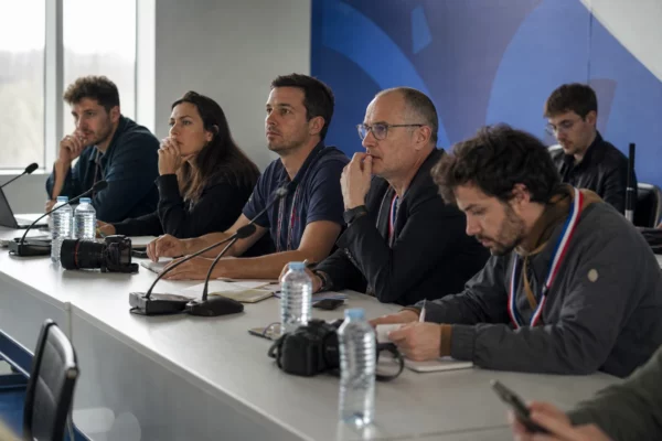 Conférence de presse du Mondial avec plusieurs journalistes anonymes, micros et carnets dans une salle lumineuse.