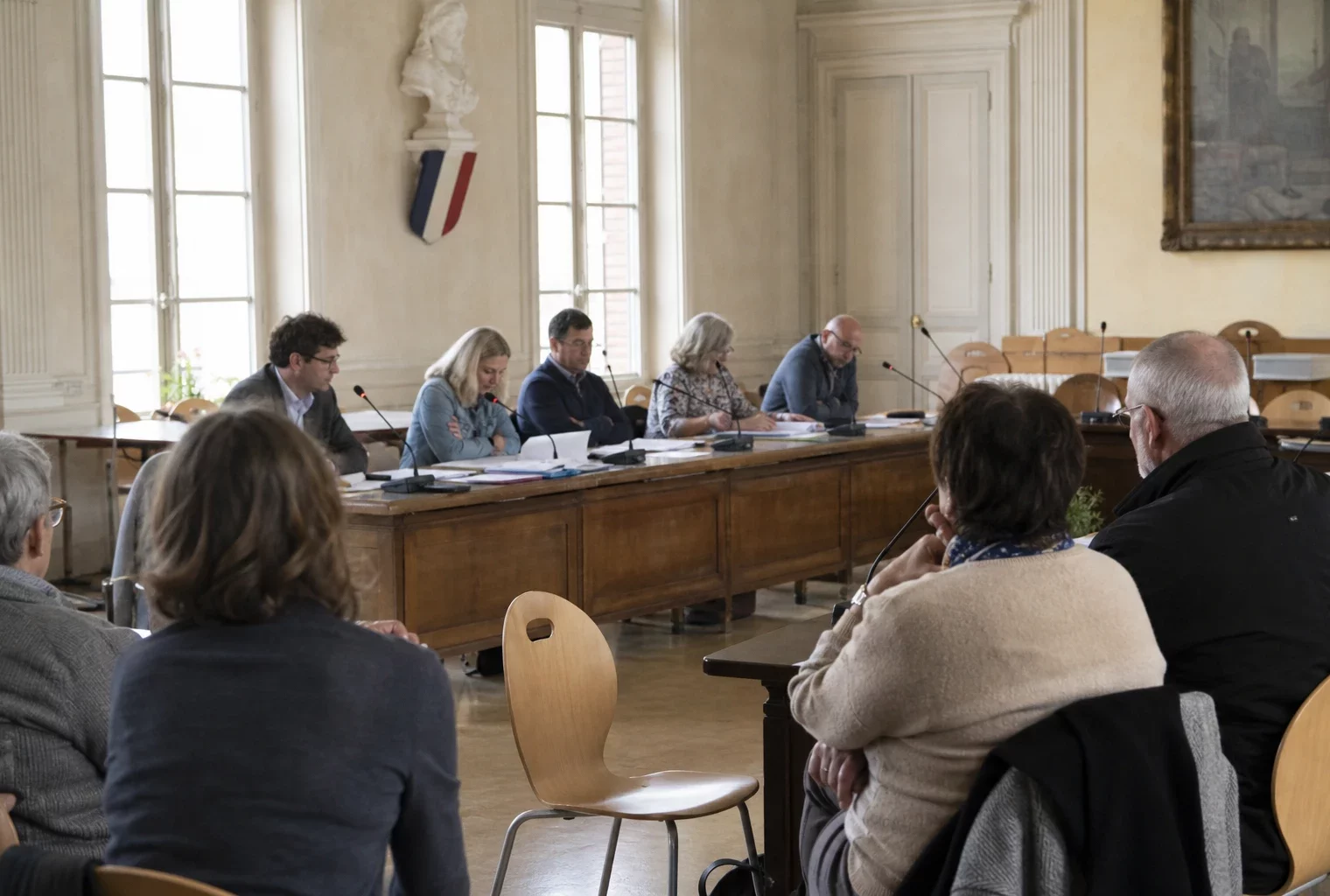 Salle municipale éclairée en daylight avec micros, dossiers flous et chaise vide lors d’un conseil local.