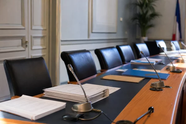 Salle municipale en plan large avec chaise vide, micros de table et dossiers flous lors d’une réunion politique.