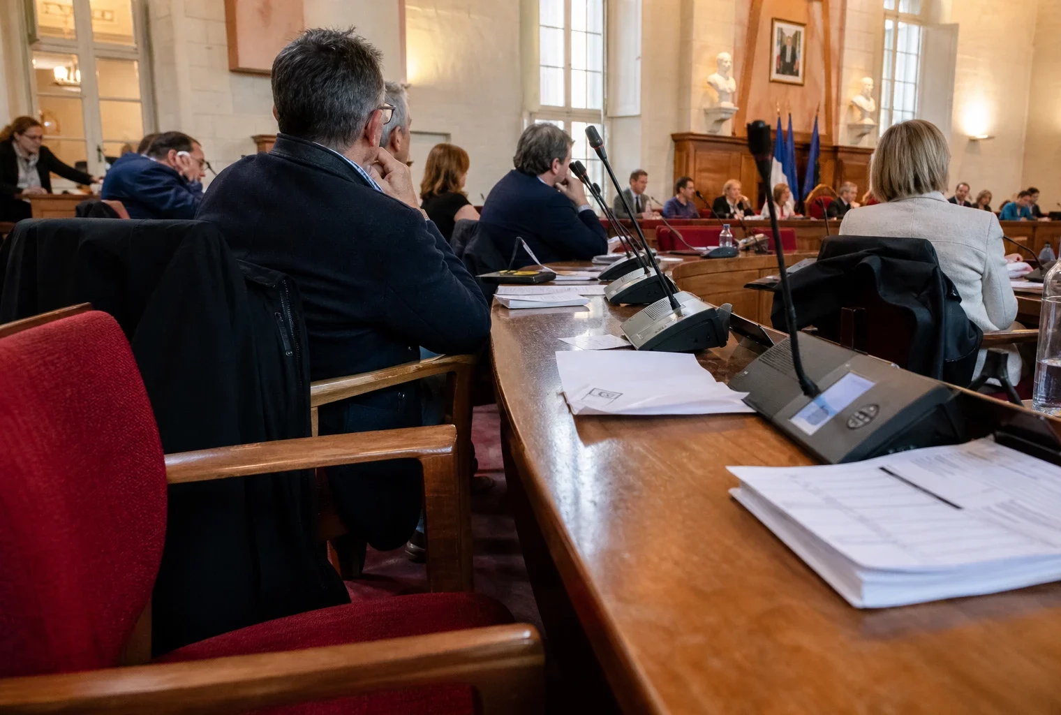 Salle municipale en réunion avec chaise vide, micros et dossiers, dans une ambiance de reportage politique.