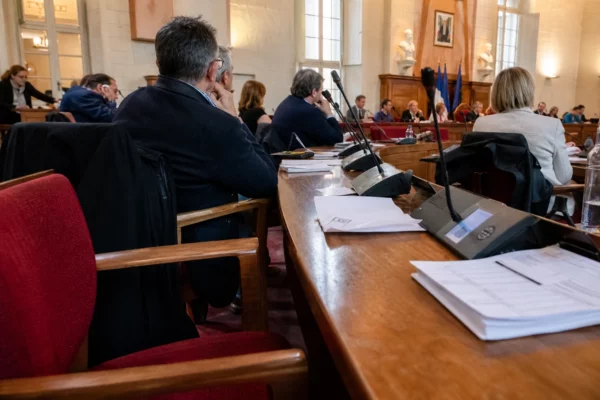 Salle municipale en réunion avec chaise vide, micros et dossiers, dans une ambiance de reportage politique.