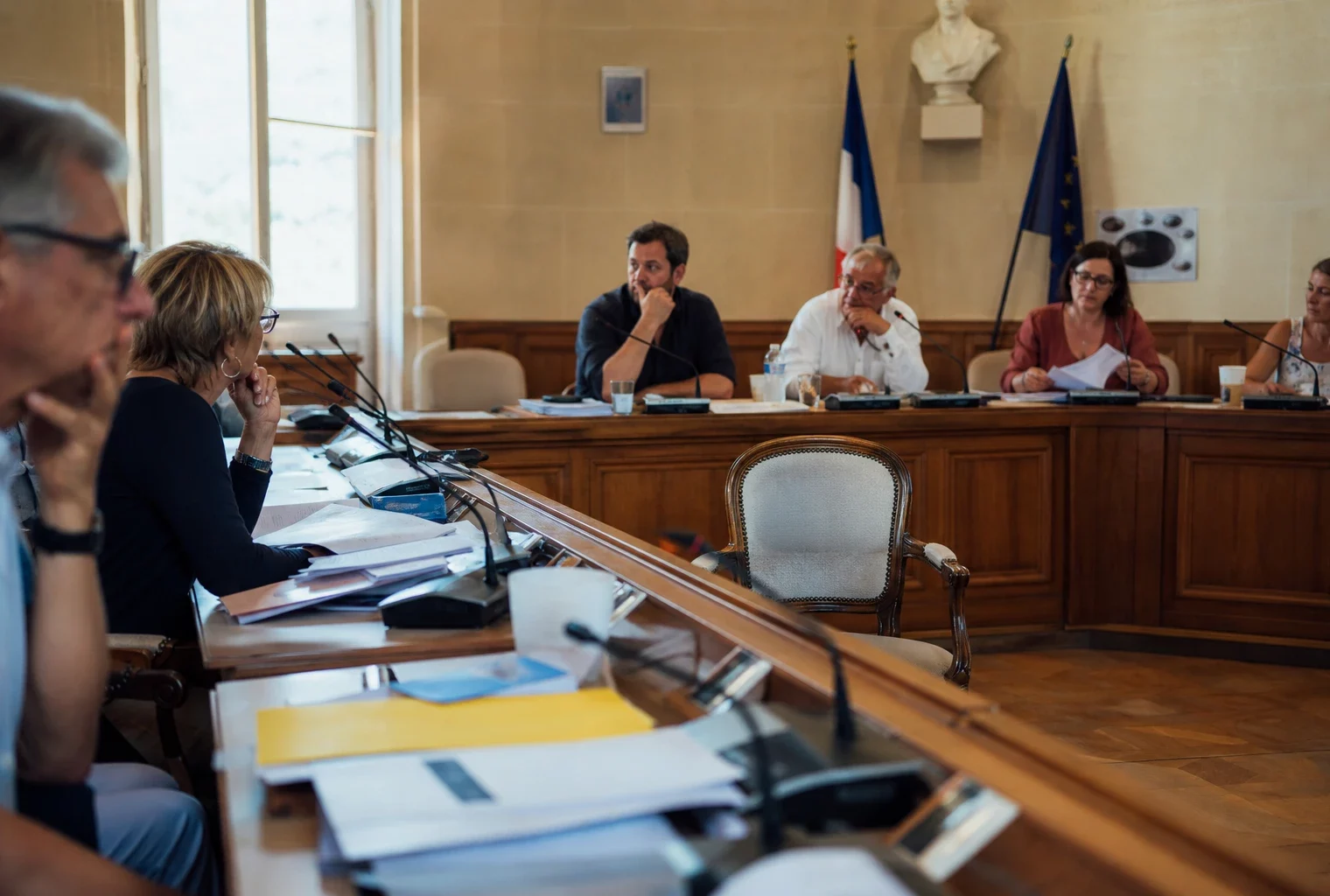 Salle municipale lumineuse avec une chaise vide, des micros et des dossiers flous lors d’une réunion politique locale.