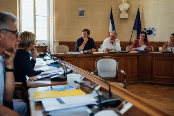 Salle municipale lumineuse avec une chaise vide, des micros et des dossiers flous lors d’une réunion politique locale.