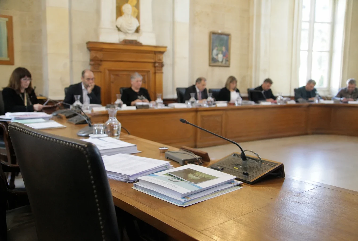 Salle municipale avec micros, dossiers flous et chaise vide lors d’une réunion sur l’eau