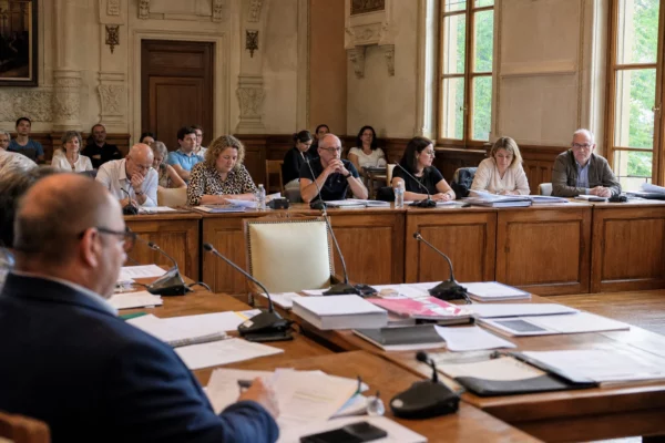 Salle municipale lumineuse avec micros, dossiers flous et chaise vide subtile lors d’un conseil local.