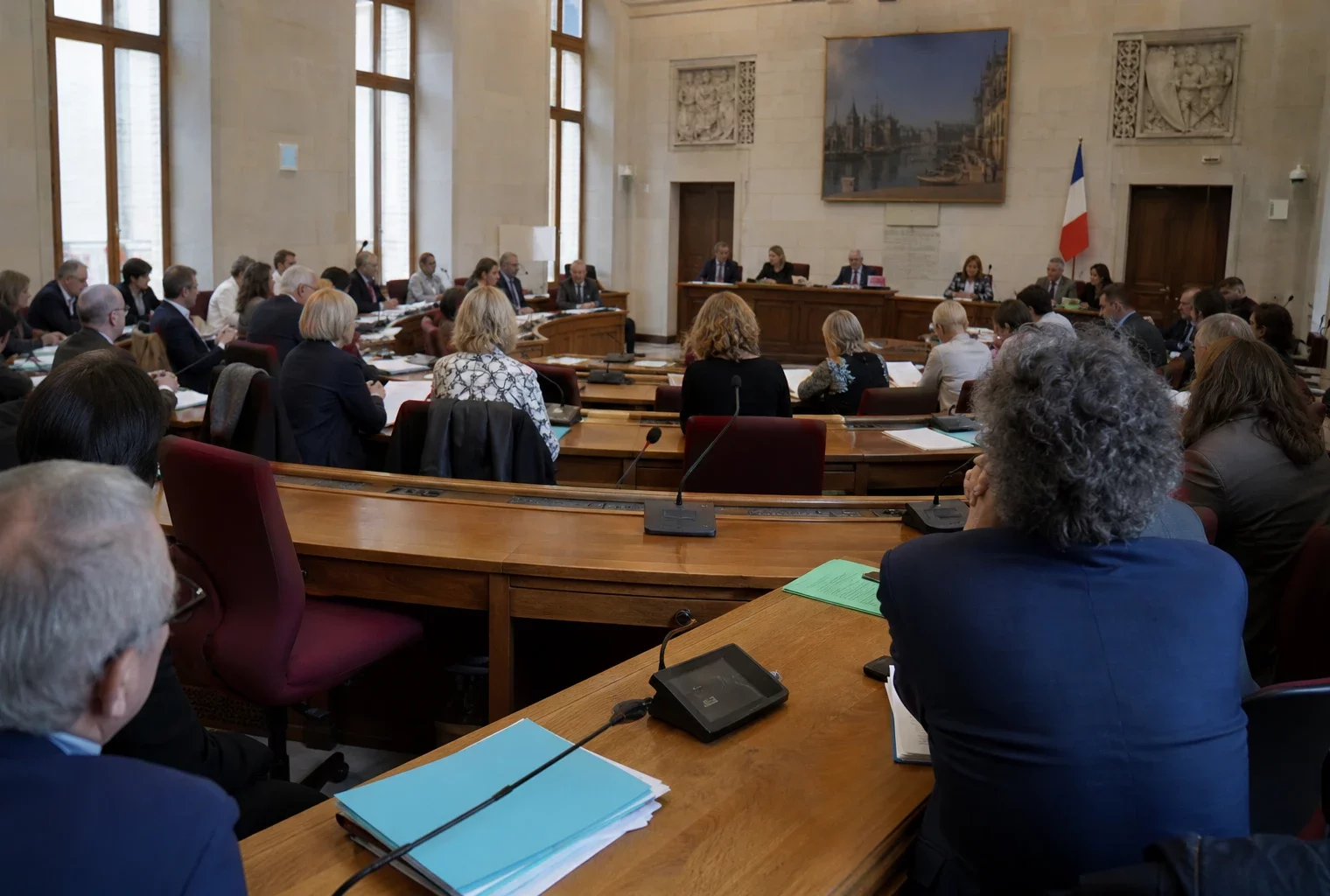 Salle du conseil municipal de Rouen, chaise vide discrète, micros et dossiers flous dans une lumière naturelle claire.