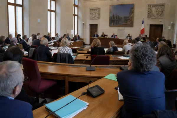 Salle du conseil municipal de Rouen, chaise vide discrète, micros et dossiers flous dans une lumière naturelle claire.
