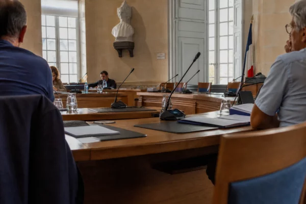 Salle de conseil municipal en lumière naturelle, avec chaise vide, micros et dossiers sur une grande table.
