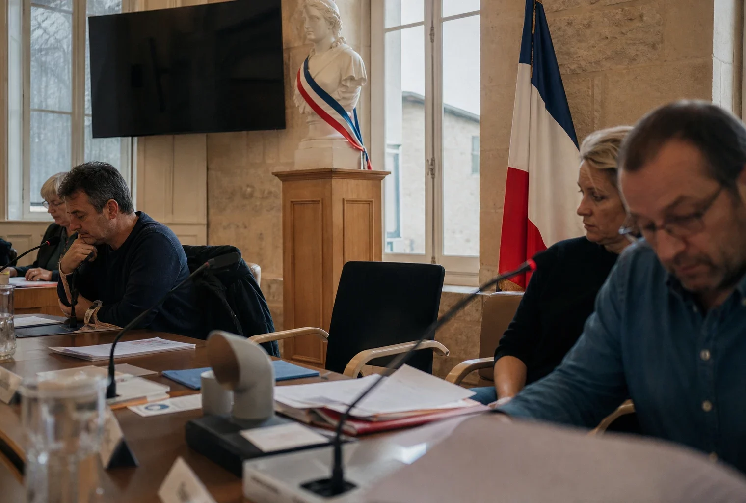 Salle municipale française avec chaise vide, micros et dossiers flous, dans une lumière naturelle claire.