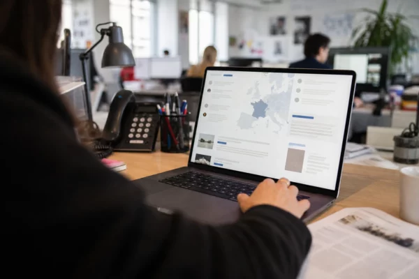 Vue en plongée légère sur un écran d’ordinateur montrant une interface abstraite, avec une main floue au premier plan dans une rédaction lumineuse.