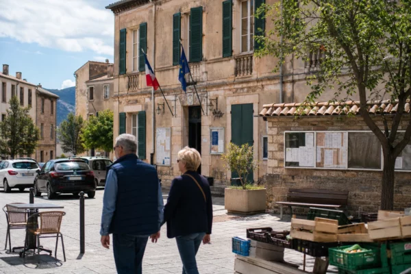Rue de ville corse près d’une mairie, avec passants anonymes et ambiance de reportage politique local.