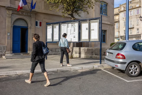 Scène de rue en Corse près d’une mairie, avec des habitants anonymes et une ambiance de reportage clair.