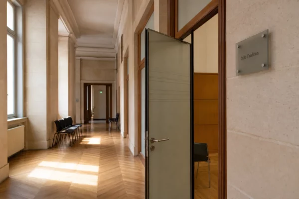 Couloir clair d’un bâtiment institutionnel français, porte d’audition entrouverte et salle vide en lumière naturelle.