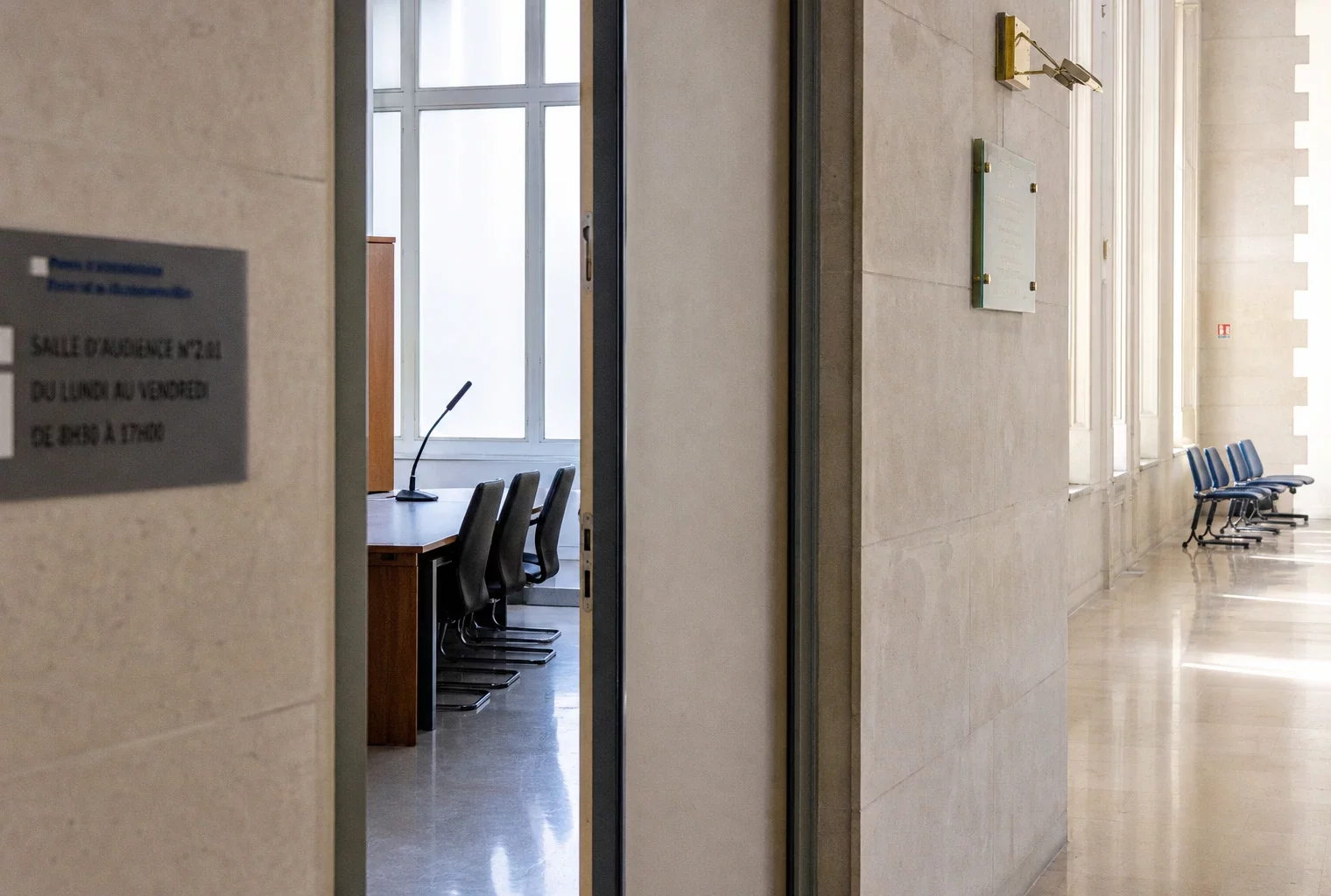 Couloir clair d’un bâtiment institutionnel français avec porte entrouverte vers une salle d’audition vide.
