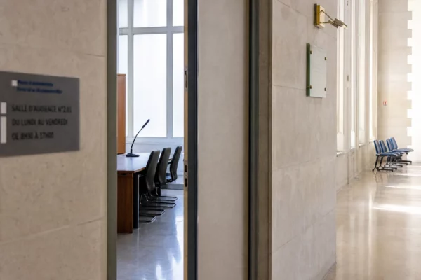Couloir clair d’un bâtiment institutionnel français avec porte entrouverte vers une salle d’audition vide.