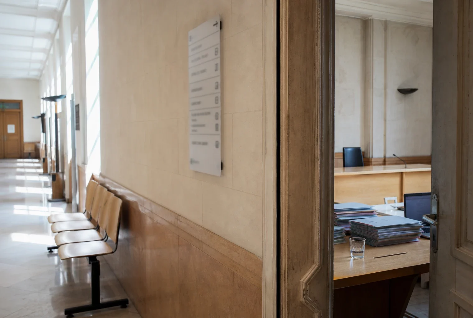 Couloir lumineux d’un bâtiment institutionnel français menant à une salle d’audition, avec portes sobres et dossiers flous.