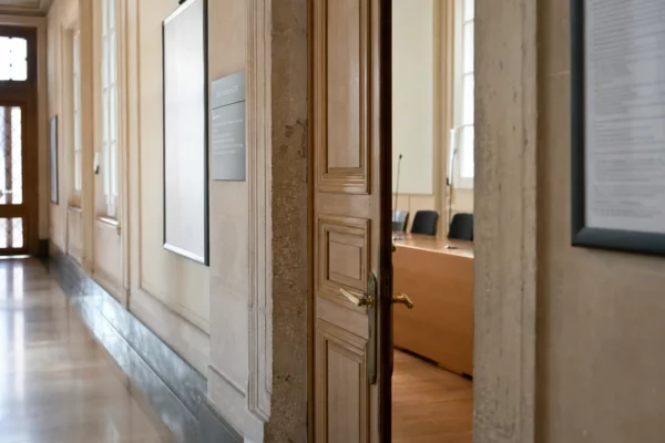 Couloir clair d’un bâtiment institutionnel français, vu en oblique vers une salle d’audition vide.