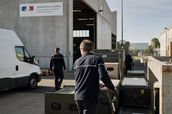 Yard of a French defense logistics site with anonymous technicians, crates, and unmarked vans in bright daylight