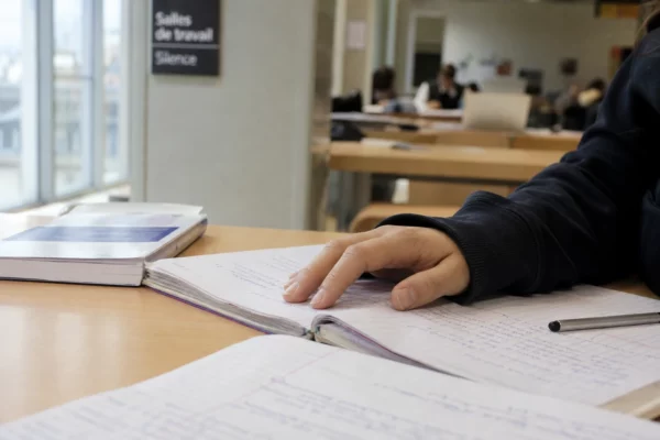 Main d’étudiant sur un cahier ouvert près d’un manuel flou dans une salle d’étude lumineuse
