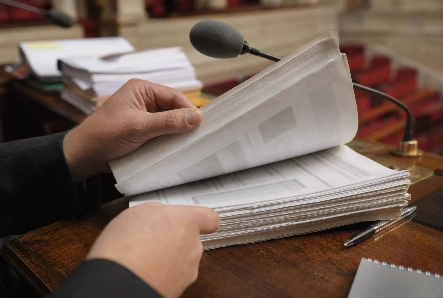 Mains anonymes feuilletant un dossier devant un micro de commission parlementaire, avec documents flous en arrière-plan.