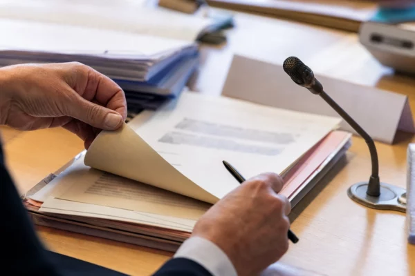 Gros plan sur un dossier d’audition parlementaire, une main tourne une page près d’un micro de commission.