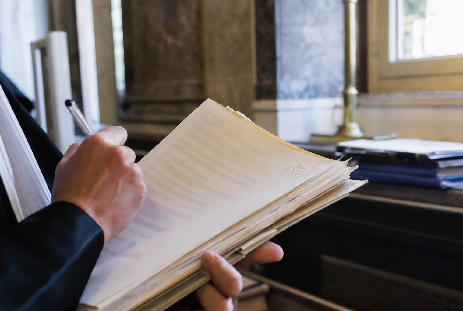 Gros plan d’une main dans un tribunal, annotant un dossier judiciaire sous une lumière naturelle claire.