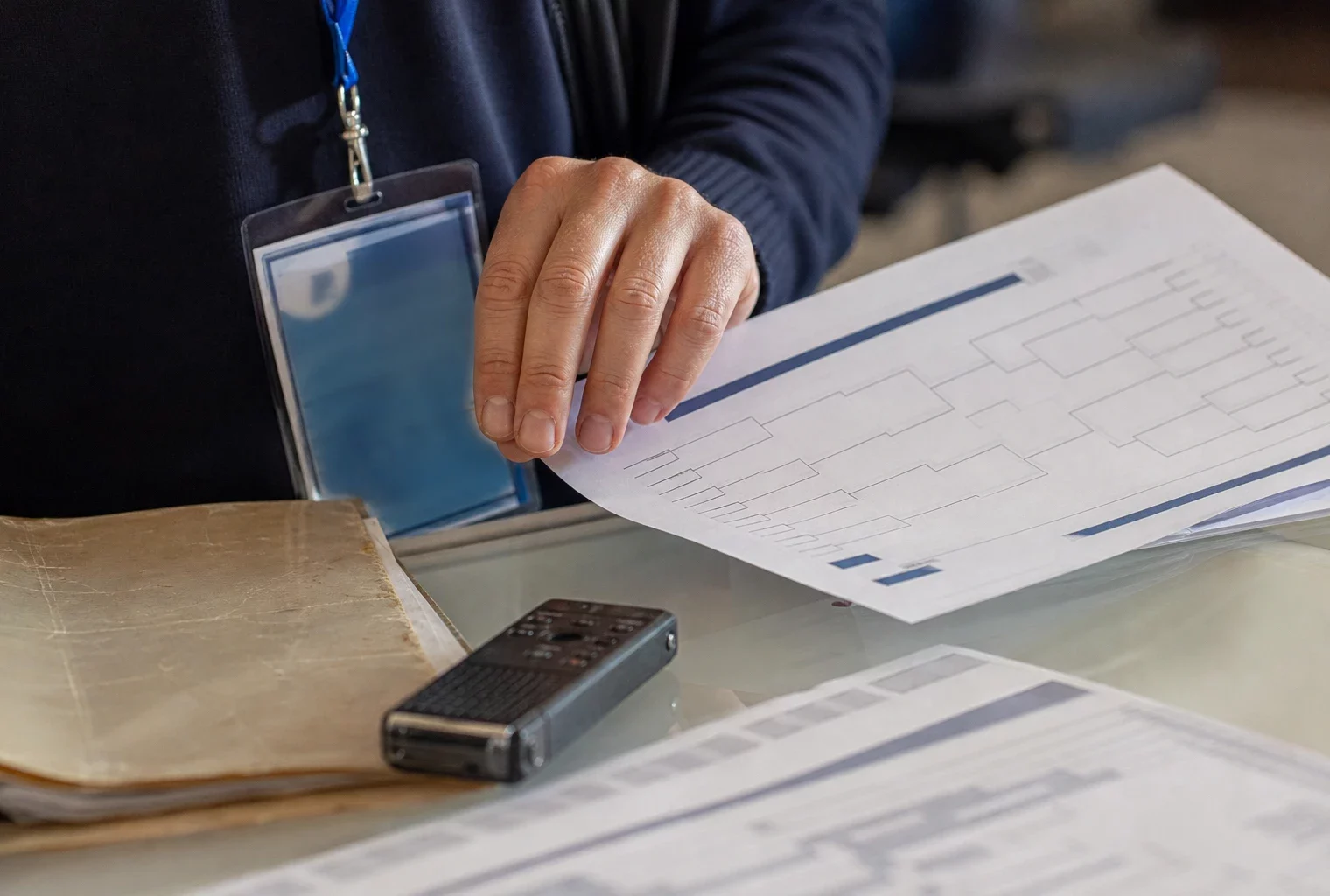 Gros plan d’une main de journaliste sur un dossier de match, avec badge presse et petit poste radio sur une table lumineuse.