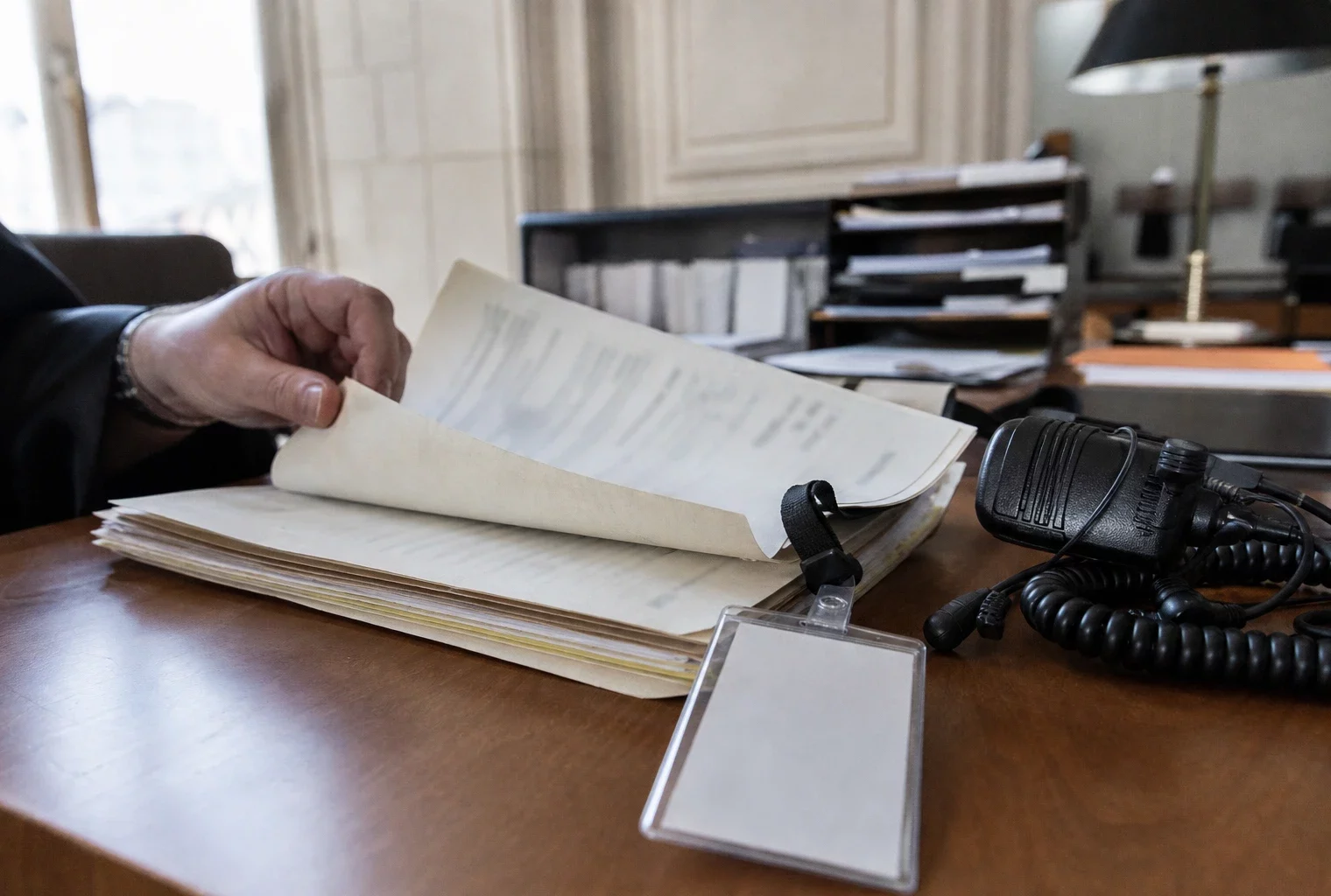 Gros plan sur un dossier de justice, un badge générique et un petit équipement radio sur un bureau éclairé par la fenêtre.