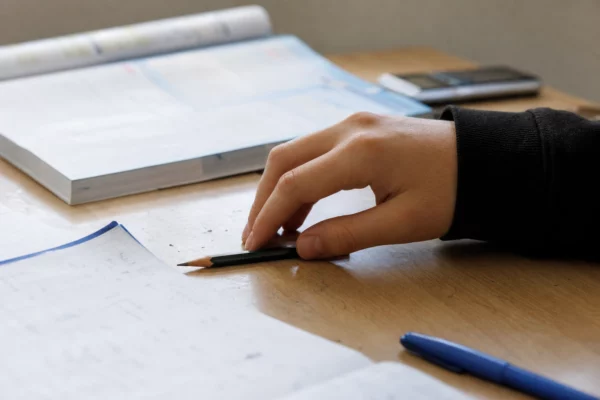 Main d’élève sur un bureau clair avec cahier de maths, stylo et manuel flou dans une salle de classe lumineuse.