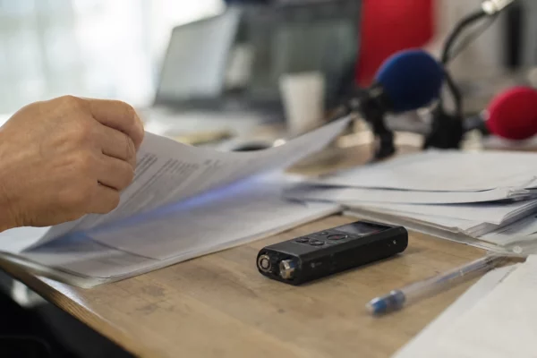 Gros plan d’une main tournant des notes sur un bureau de rédaction, avec enregistreur audio et papiers flous.
