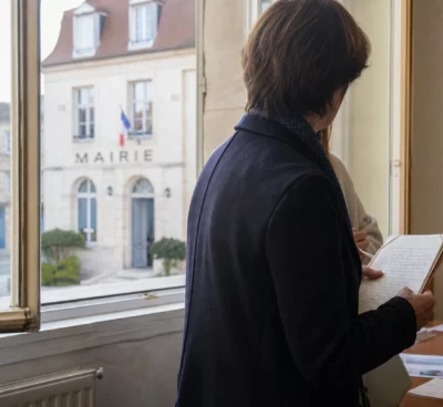 Collaborateur municipal anonyme tenant un dossier dans un couloir de mairie, avec une rue française floue en arrière-plan.