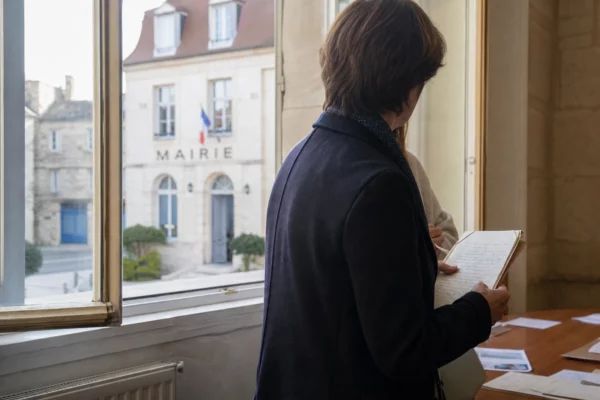 Collaborateur municipal anonyme tenant un dossier dans un couloir de mairie, avec une rue française floue en arrière-plan.