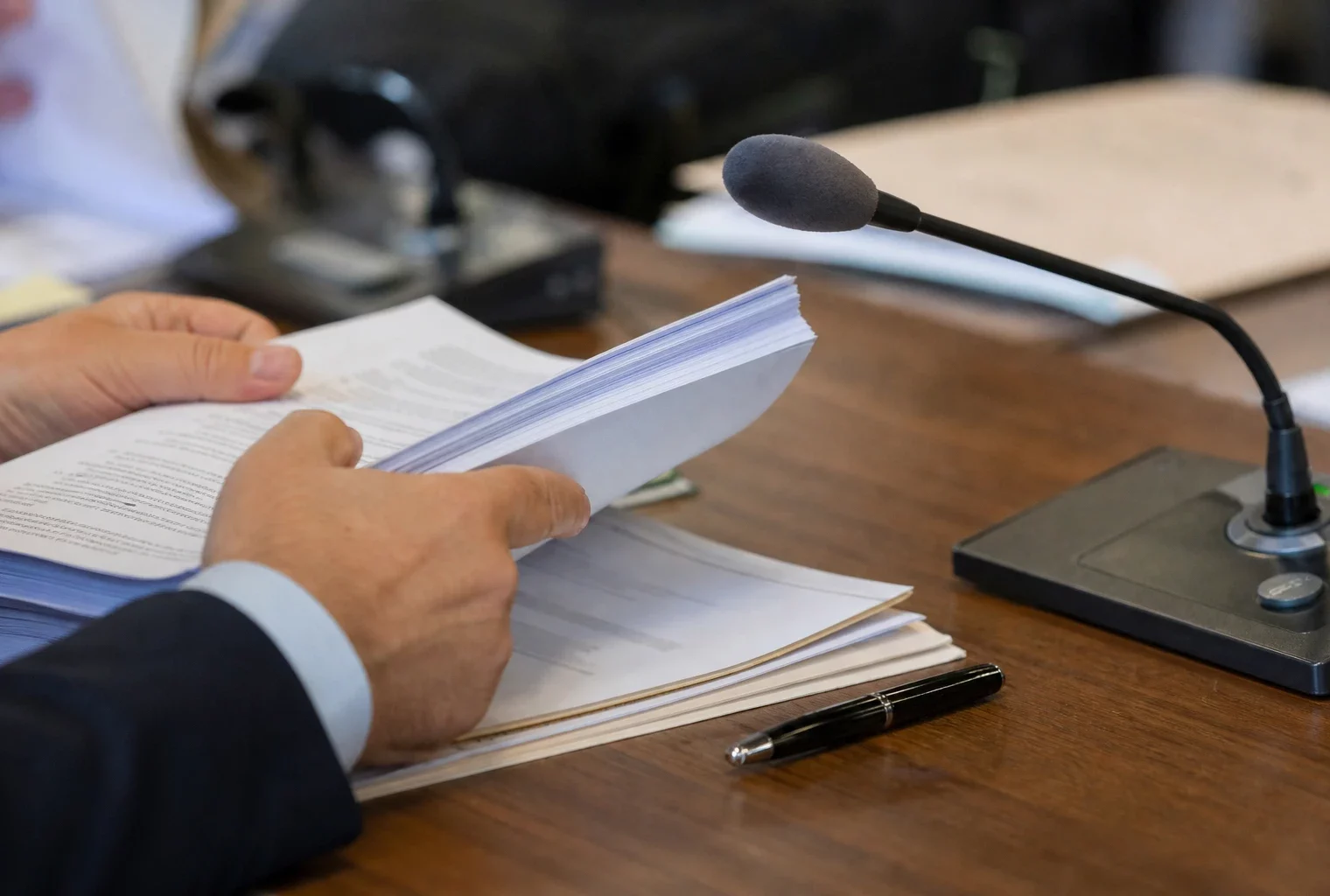 Mains anonymes, micro de commission et dossier flou dans une salle d’audition française éclairée par la fenêtre.