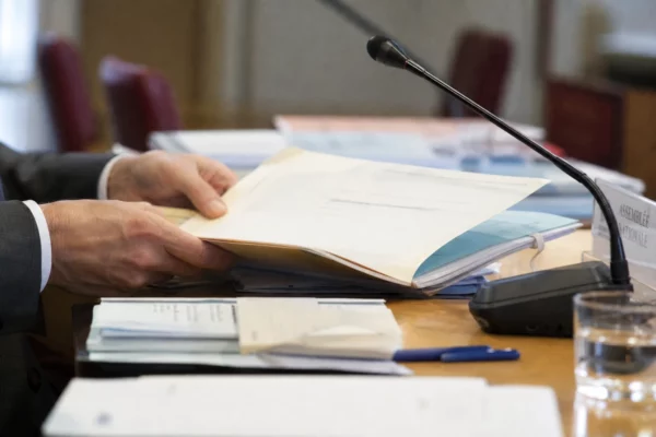 Gros plan sur des mains anonymes, un dossier et un micro de commission dans un cadre institutionnel français.