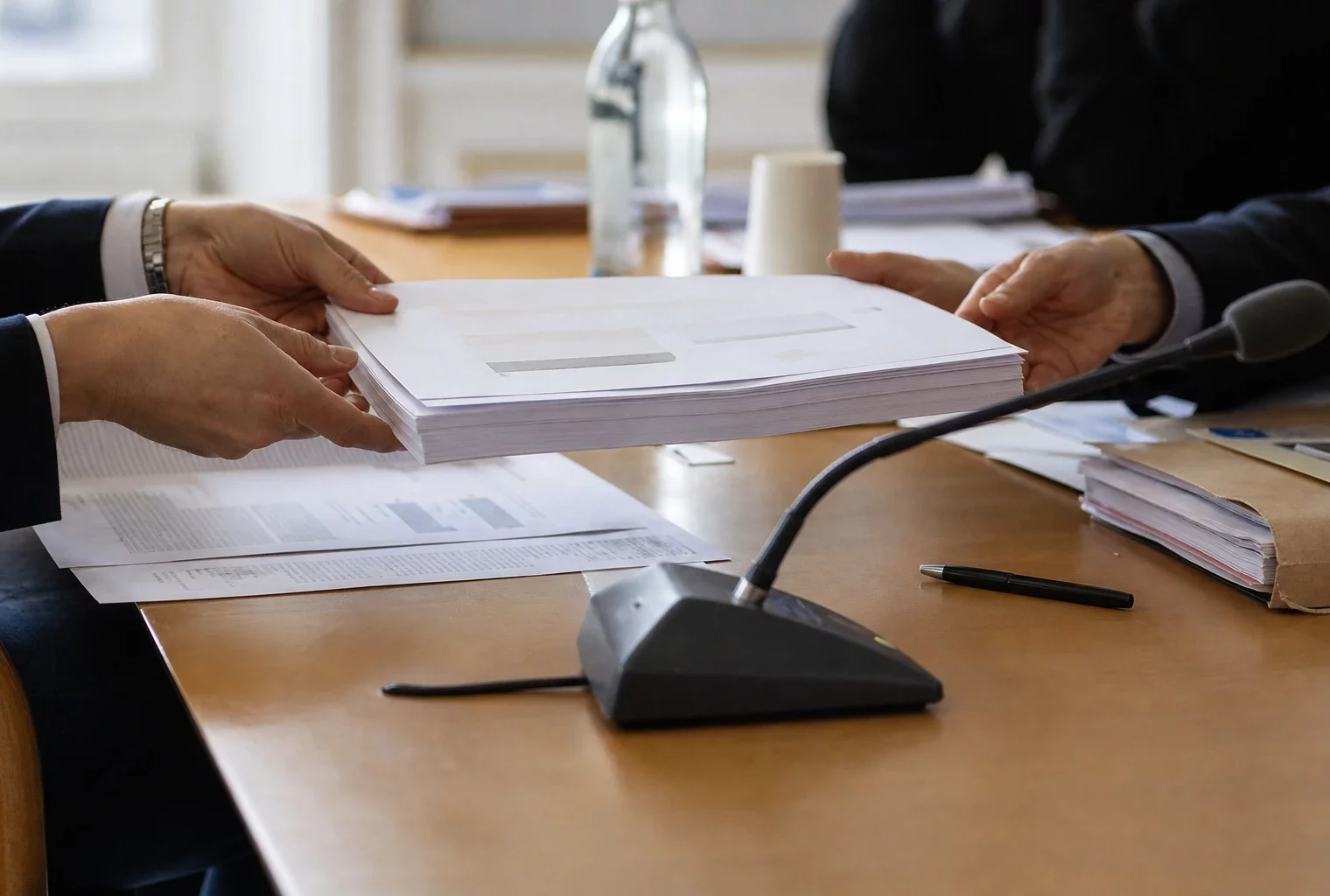 Dossier de commission et micro de réunion, mains anonymes et documents flous dans une salle institutionnelle française.
