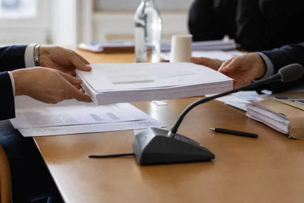 Dossier de commission et micro de réunion, mains anonymes et documents flous dans une salle institutionnelle française.