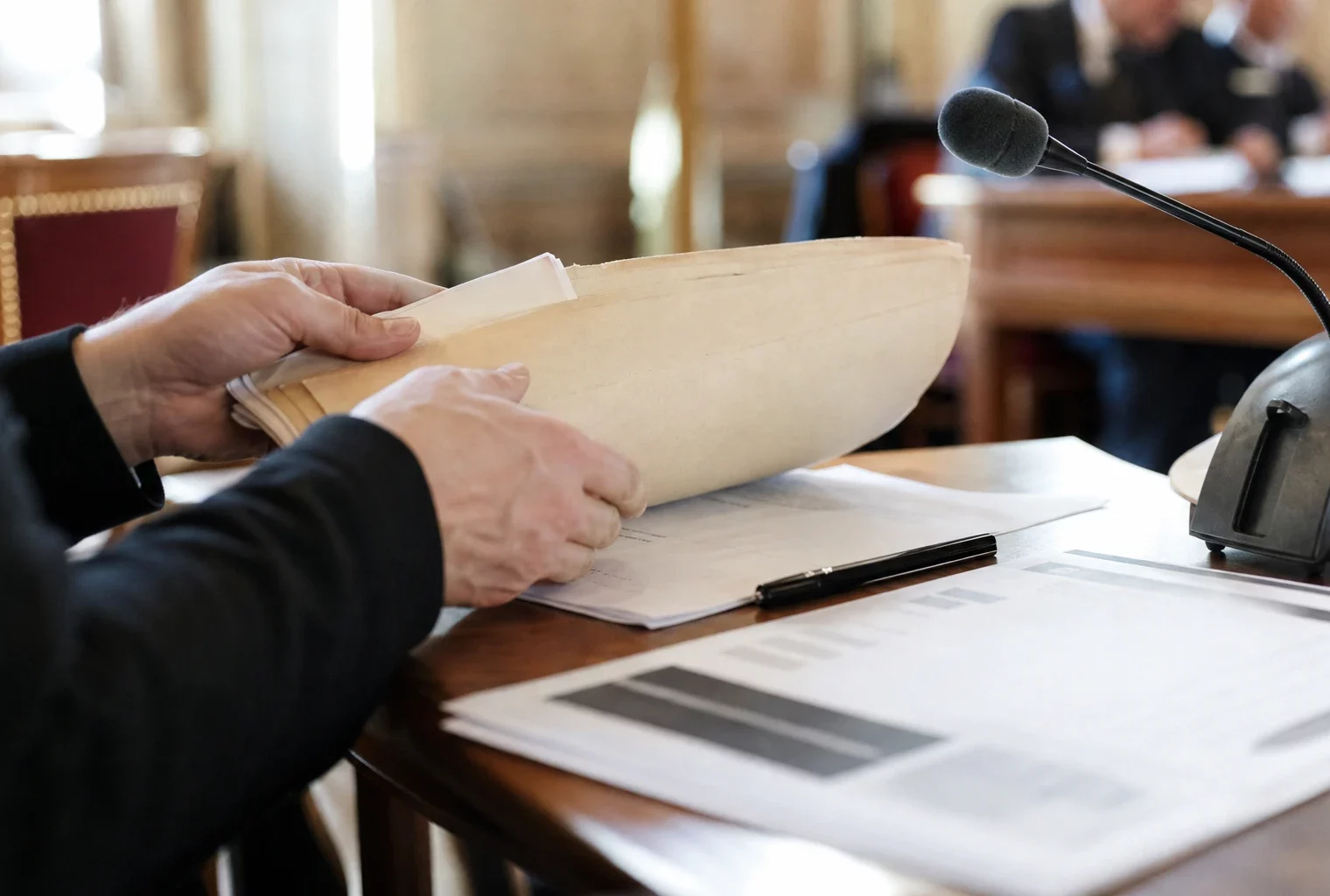 Mains anonymes feuilletant un dossier lors d’une audition, avec micro de commission et documents flous