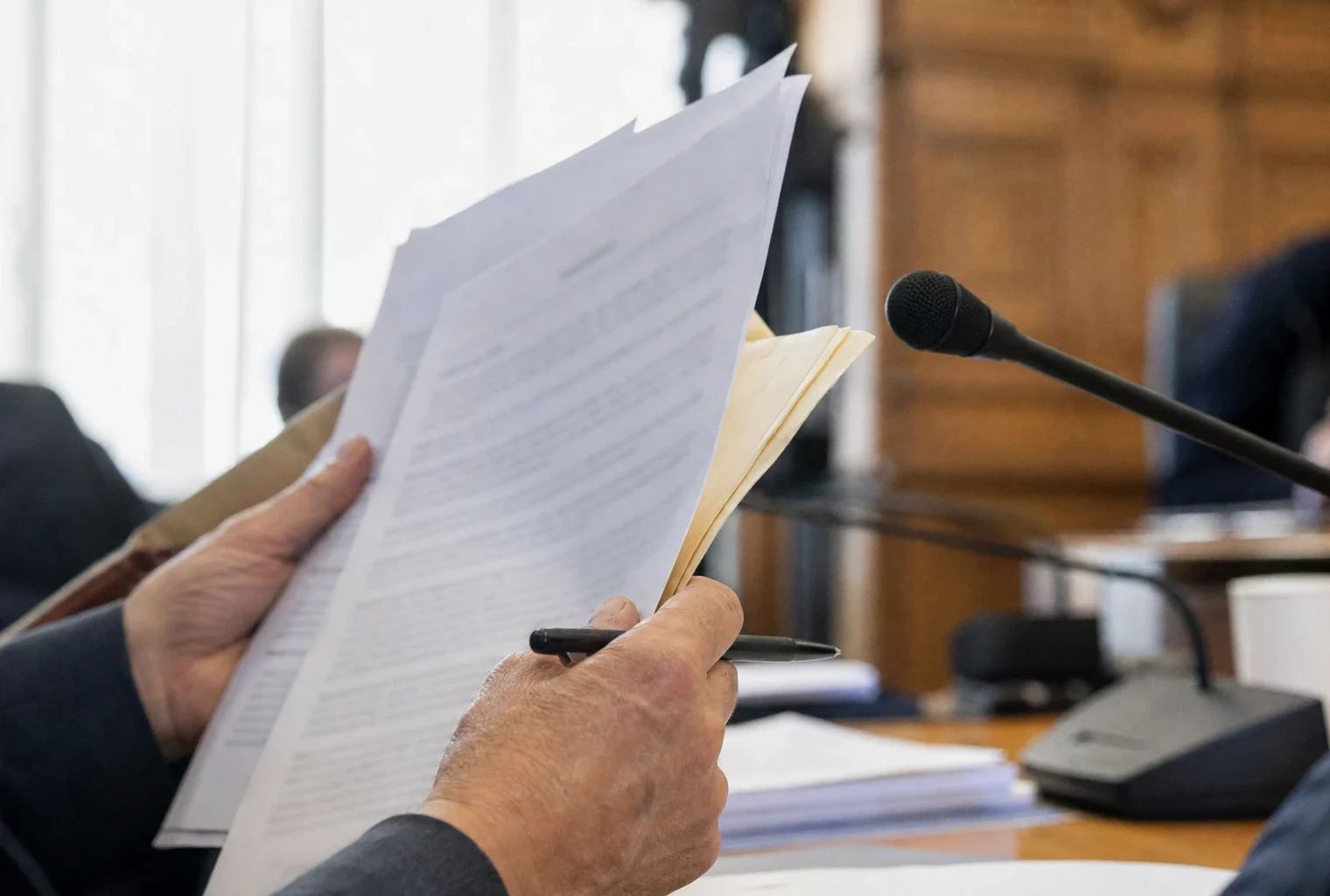 Mains anonymes avec dossier et micro de commission lors d’une audition parlementaire, en lumière naturelle.