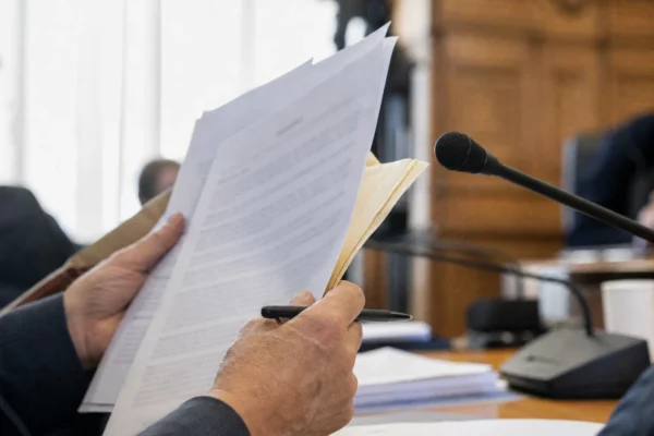 Mains anonymes avec dossier et micro de commission lors d’une audition parlementaire, en lumière naturelle.