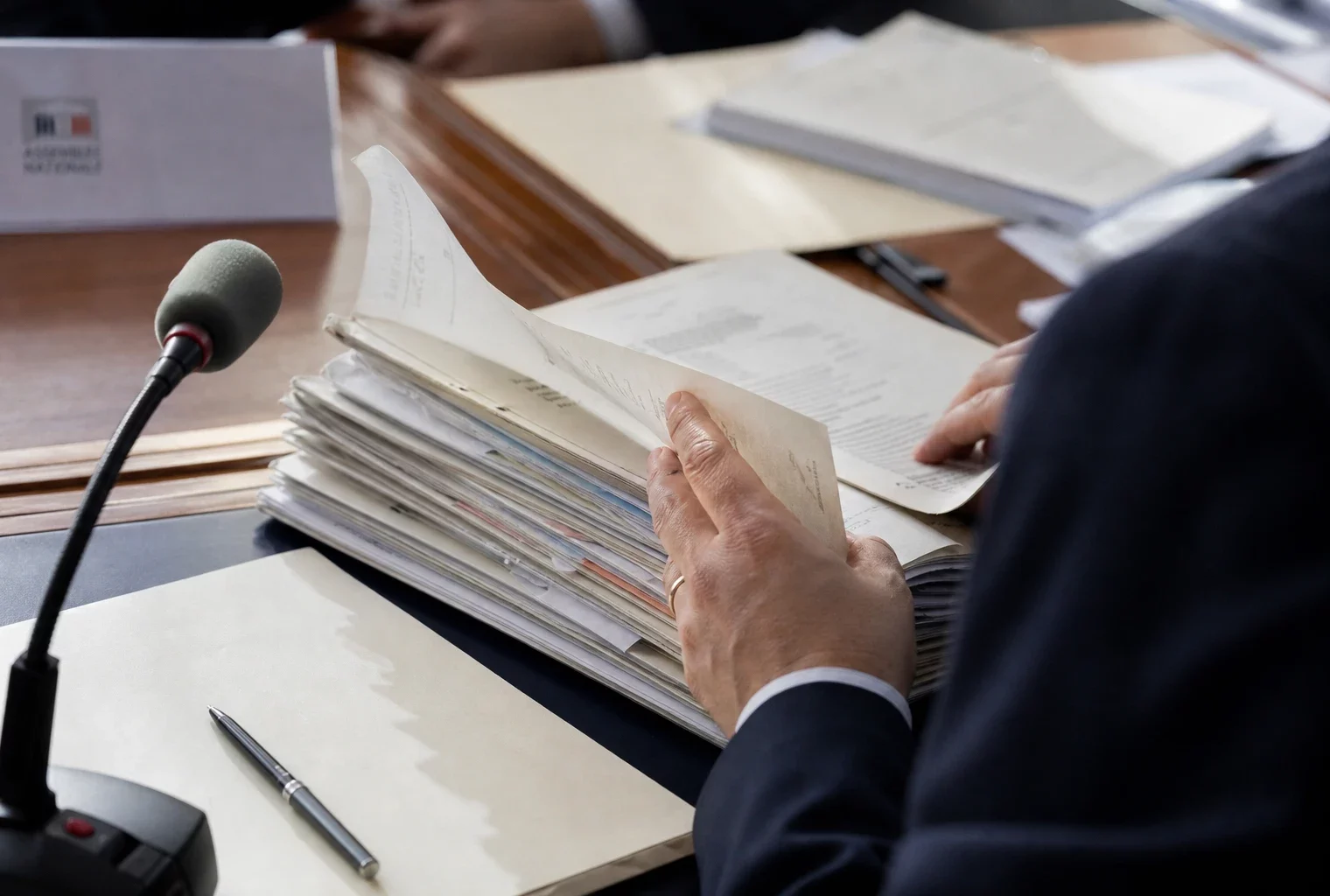 Main anonyme feuilletant un dossier devant un micro de commission dans une salle de justice lumineuse.