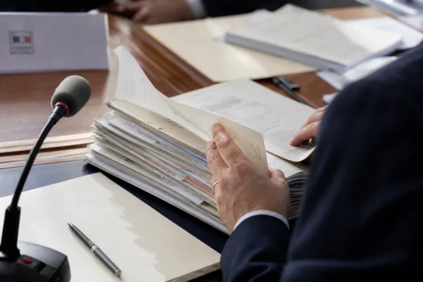 Main anonyme feuilletant un dossier devant un micro de commission dans une salle de justice lumineuse.