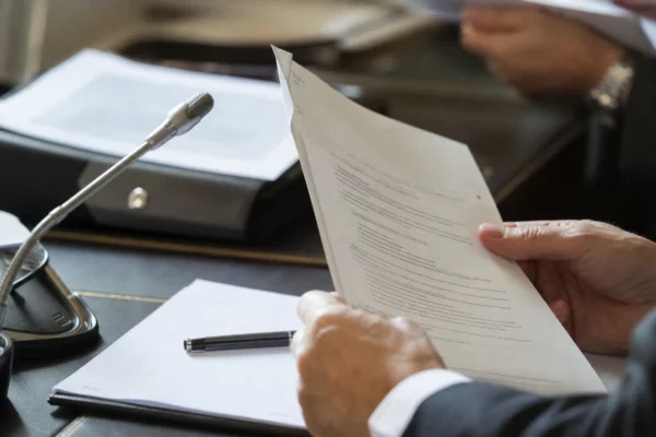 Mains anonymes consultant un dossier devant un micro de commission parlementaire, dans une lumière naturelle claire.