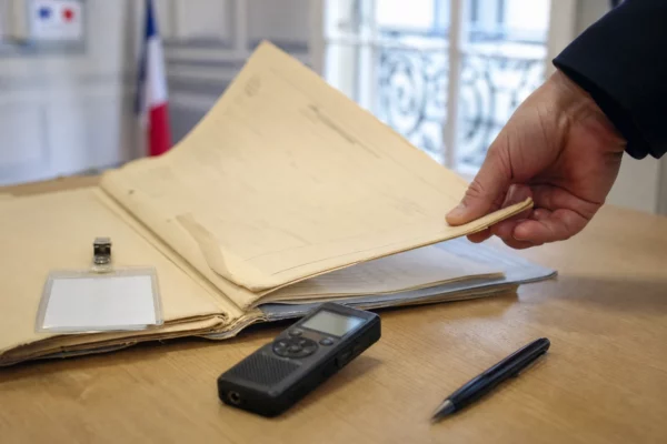Gros plan d’un dossier administratif, d’un badge neutre et d’un enregistreur sur une table éclairée par une fenêtre.