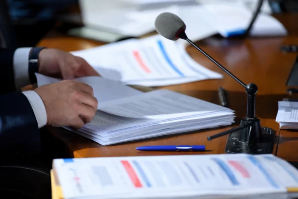 Mains anonymes feuilletant un dossier de commission près d’un micro, dans une salle parlementaire lumineuse.