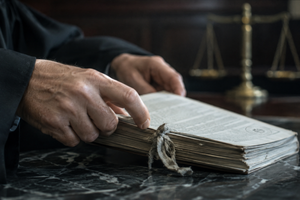 Mains en robe judiciaire ouvrant un ancien dossier juridique sur un bureau en marbre, avec une balance de justice floue à l’arrière-plan.