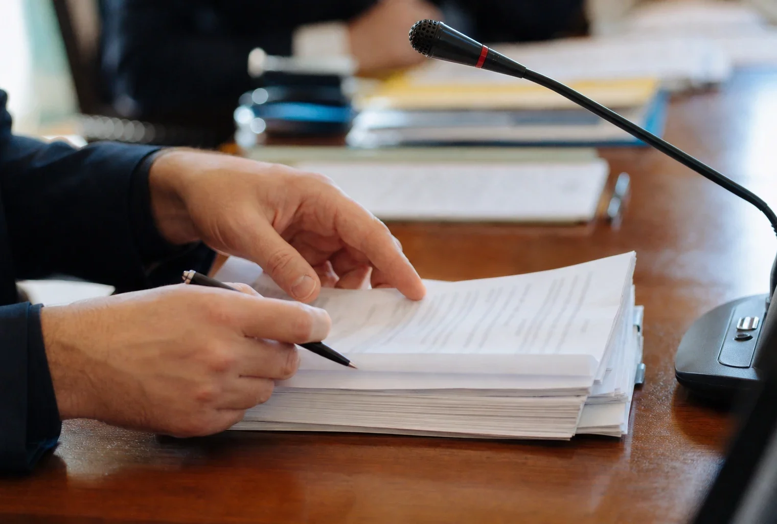 Mains anonymes manipulant un dossier de nomination près d’un micro de commission, en lumière naturelle.