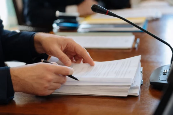 Mains anonymes manipulant un dossier de nomination près d’un micro de commission, en lumière naturelle.