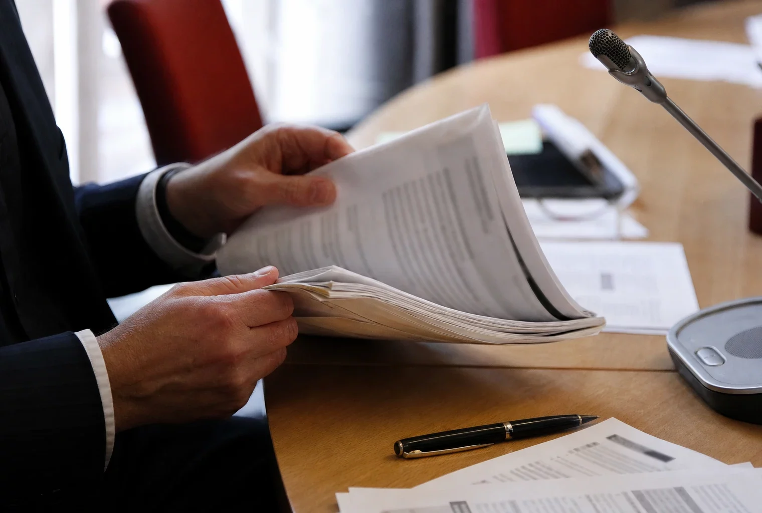 Mains anonymes feuilletant un dossier de nomination près d’un micro de commission parlementaire, avec documents flous.