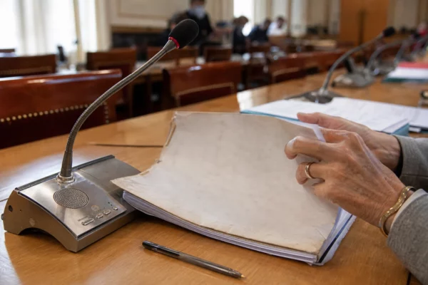 Dossier de nomination manipulé lors d’une audition, avec micro de commission et documents flous dans une salle institutionnelle.